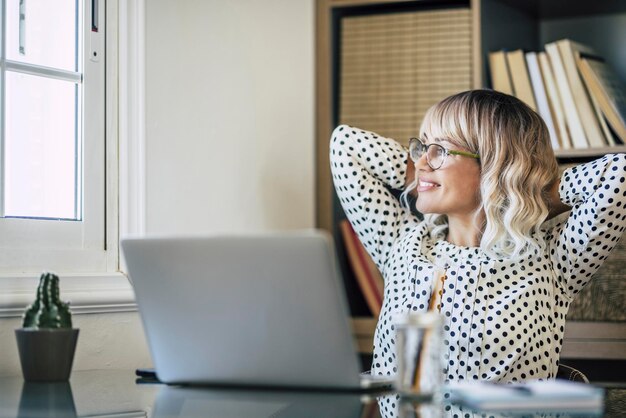 Mulheres trabalhadoras relaxadas esticando o ombro e olhando pela janela satisfeitas e felizes empresária moderna sorrindo após o dia de trabalho com o laptop no escritório doméstico