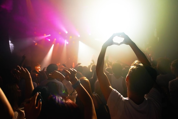 Multidão em música de concerto e assistir festival de música de performance de banda luz e energia com visão traseira do público As pessoas dançam com as mãos em forma de coração músico no palco e festival na Flórida