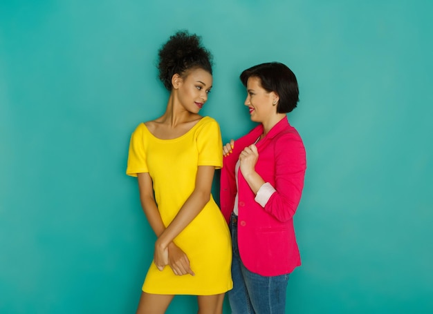 Multiethnische Freundschaft. Charmante schwarze und kaukasische Mädchen in farbenfroher Freizeitkleidung posieren auf blauem Studiohintergrund mit Kopienraum, die miteinander reden und sich ansehen