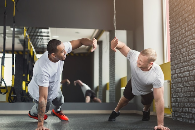 Multiethnische glückliche Männer trainieren im Fitnessclub. Fröhliche Afroamerikaner und Kaukasier, die Paarplanken oder Liegestütze machen, sich gegenseitig High-Five geben, zusammen trainieren, Platz kopieren