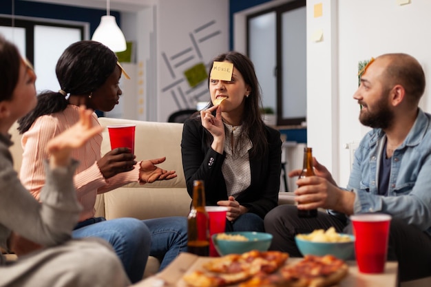 Multiethnische Gruppe von Freunden, die Haftnotizen verwenden, um zu erraten, wer nach Feierabend auf einer Büroparty zusammen spielt. Geschäftsleute spielen Scharaden, essen Pizza und trinken Bier nach der Arbeit.