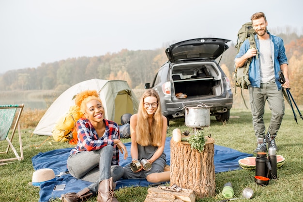 Multiethnische Gruppe von Freunden, die lässig gekleidet ein Picknick während der Erholung im Freien mit Zelt, Auto und Wanderausrüstung in der Nähe des Sees machen