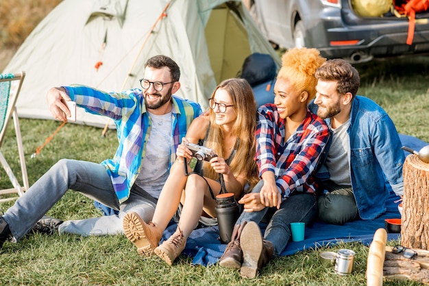 Multiethnische Gruppe von Freunden, die lässig gekleidet sind und Spaß haben, während der Erholung im Freien mit Zelt, Auto und Wanderausrüstung in der Nähe des Sees zusammen ein Selfie-Foto zu machen