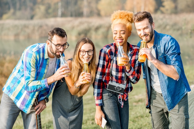 Multiethnische Gruppe von Freunden, die lässig gekleidet sind und Spaß mit Getränken während der Erholung im Freien auf dem Campingplatz in der Nähe des Waldes haben