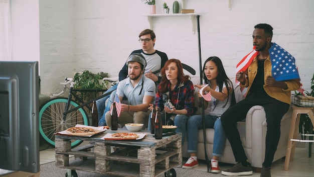 Multiethnische Gruppe von Freunden, Sportfans mit US-Nationalflaggen, die Hockey-Meisterschaften im Fernsehen verfolgen