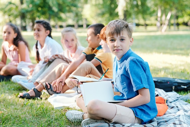 Multiethnische gruppe von kindern, die auf grünem gras sitzen und modellplaneten halten, während sie den astronomieunterricht im freien im sonnenlicht genießen