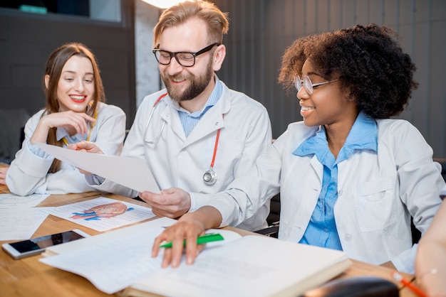 Multiethnische Gruppe von Medizinstudenten in Uniform, die eine Diskussion führen, die zusammen am Schreibtisch mit verschiedenen medizinischen Sachen im Klassenzimmer sitzt