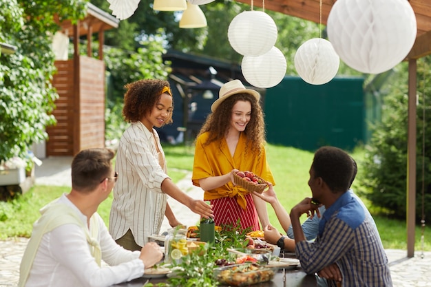 Multiethnische Gruppe von Menschen, die Abendessen auf der Außenterrasse im Sommer genießen, konzentrieren sich auf lächelnde junge Frau, die frisches Obst und Beeren über Tisch reicht