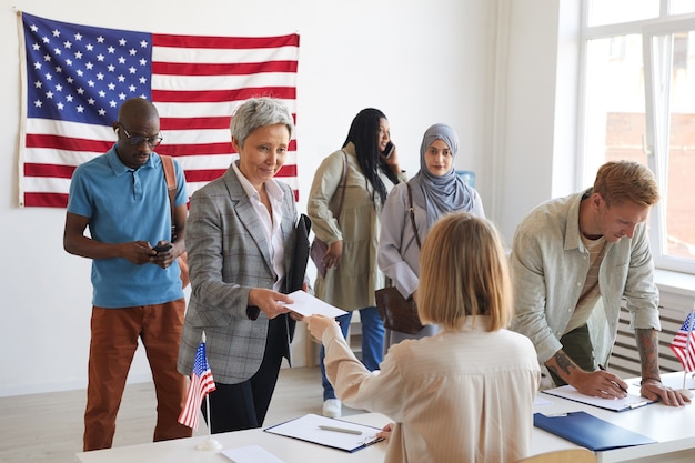 Multiethnische Gruppe von Menschen, die sich am Wahltag im Wahllokal mit amerikanischen Flaggen registrieren lassen, Kopierraum