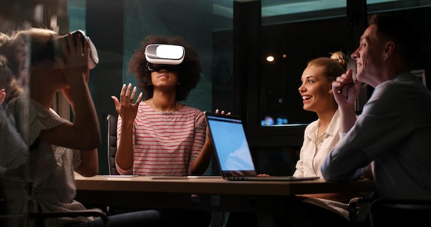 Multiethnisches Geschäftsteam mit Virtual-Reality-Headset in Nachtbürositzung Entwicklertreffen mit Virtual-Reality-Simulator am Tisch im Kreativbüro.
