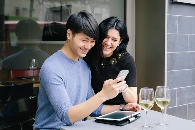 Multiethnisches Paar in einem Restaurant, das mit einem Glas Weißwein auf das Telefon schaut