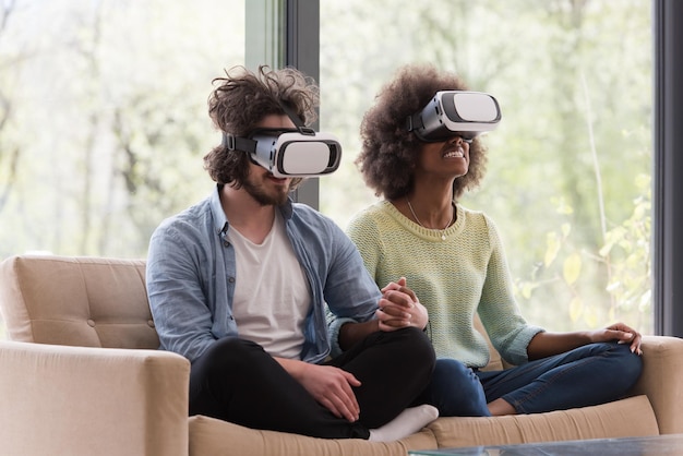 Multiethnisches Paar mit Virtual-Reality-Headset im Wohnzimmer zu Hause, Leute, die mit neuer Trendtechnologie spielen