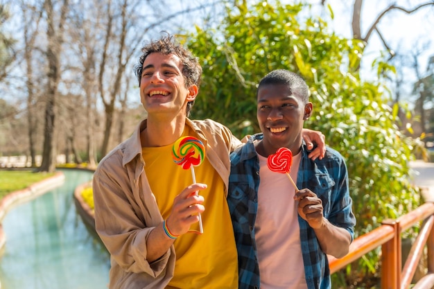 Multiethnisches schwules männliches Paar, das ein Lollipop-lgbt-Konzept isst, das Spaß hat und in einem Park lächelt