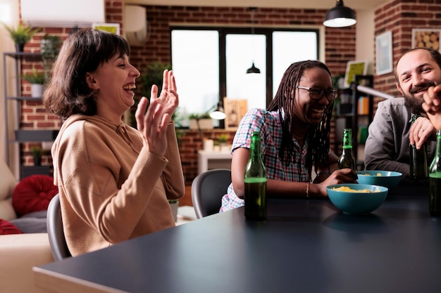 Multikulturelle Gruppe von Freunden, die zu Hause im Wohnzimmer sitzen und zusammen lachen und scherzen. Multiethnische junge Erwachsene haben Spaß, während sie Snacks und Getränke genießen, während sie am Tisch sitzen.