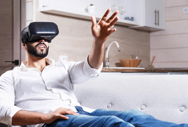 Multikultureller bärtiger Mann, der auf dem Sofa liegt und in der modernen Wohnung eine Virtual-Reality-Brille verwendet