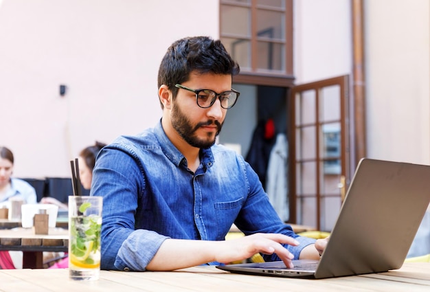 Multikultureller intelligenter Student im Jeanshemd, der etwas am Laptop durchstöbert, während er im Café sitzt