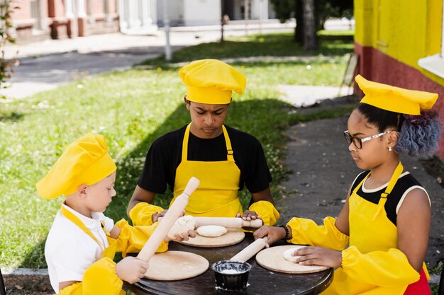 Multirassische Köche Kinder in gelber Kochmütze und Schürze, die Teig für die Bäckerei kochen Schwarzafrikanisches und kaukasisches Kind, das zusammen kocht und Spaß hat