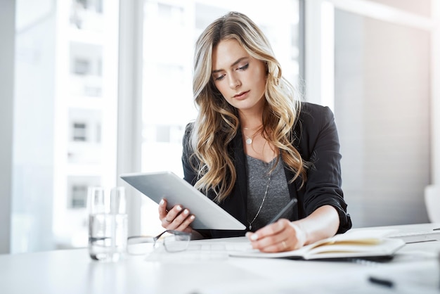 Multitarefa faz acontecer Foto de uma jovem empresária usando um tablet digital e escrevendo notas em sua mesa em um escritório moderno