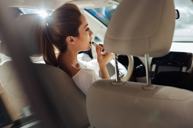 Multitarefa perigosa. Mulher jovem e bonita atraente sentada ao volante e dirigindo o carro enquanto se maquia