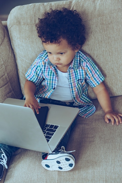 Multitasking-Baby. Blick aus der Vogelperspektive auf den kleinen afrikanischen Jungen, der seinen Laptop und sein Smartphone benutzt, während er zu Hause auf der Couch sitzt