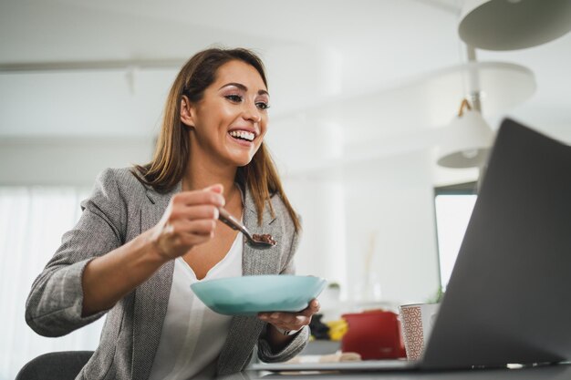 Multitasking-junge Geschäftsfrau, die während einer Pandemie frühstückt und Laptop benutzt, während sie zu Hause arbeitet.