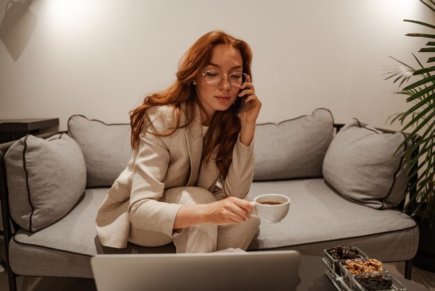Multitasking-Konzept. Arbeit zu Hause, Geschäftsreise. Eine sehr beschäftigte rothaarige junge Frau in einem stylischen Business-Anzug und am Telefon benutzt Papiere und einen Laptop.