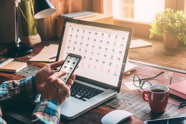 Foto multitasking. nahaufnahme eines mannes, der ein smartphone hält, während er am rustikalen holztisch mit laptop sitzt