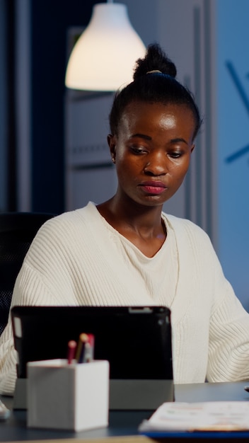 Multitasking schwarze Geschäftsfrau, die gleichzeitig an Laptop und Tablet arbeitet und Überstunden im Start-up-Büro macht. Beschäftigter afrikanischer Mitarbeiter, der Finanzstatistiken analysiert, das Schreiben überfordert, sucht.