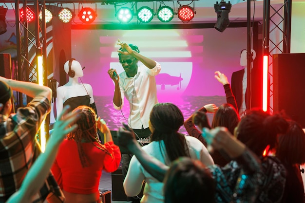 Multitud bailando y levantando las manos mientras el músico actúa en el escenario del club. Gente moviéndose en la pista de baile y divirtiéndose mientras disfruta de un concierto de música electrónica en vivo en una discoteca