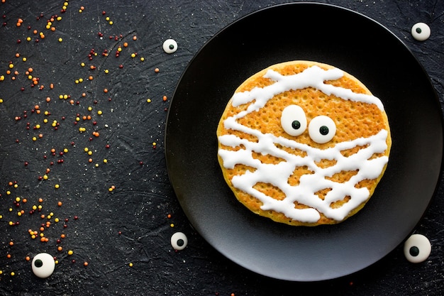 Mumienpfannkuchen lustige Idee für Kinder zum Halloween-Frühstück Kürbispfannkuchen mit cremefarbenen und süßen Augen formten Mumie auf schwarzem Teller von oben