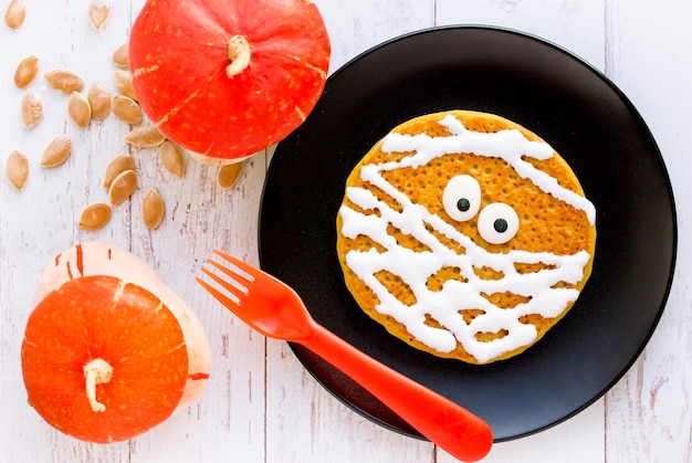 Mumienpfannkuchen lustige Idee für Kinder zum Halloween-Frühstück Kürbispfannkuchen mit cremefarbenen und süßen Augen formten Mumie auf schwarzem Teller von oben