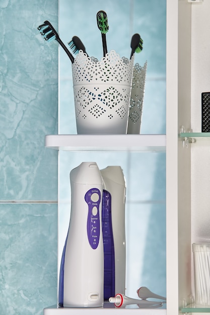 Munddusche oder Wasserflosser mit austauschbarer Düse und Zahnbürsten unter den Toilettenartikeln auf dem Wandschrankregal im Badezimmer.