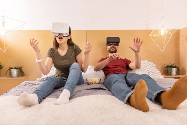 Foto mundo sin límites. alegre pareja complacida asombrada usando gafas vr mientras se relaja en la cama y saborea palomitas de maíz