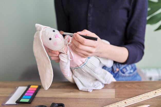 Muñeca de conejito hecha a mano en manos de una adolescente, niña pinta sus mejillas con cosméticos. Pasatiempos, ocio, creatividad y habilidades adolescentes.