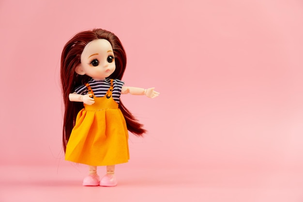 Foto una muñeca de juguete infantil con cabello oscuro y un vestido naranja de pie sobre un fondo rosa. un juguete de plástico para niños. una muñeca de plástico indica un espacio vacío para un diseño o espacio de copia de texto.