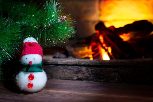 Muñeco de nieve debajo de un árbol de Navidad cerca de una chimenea ardiente, primer plano, composición navideña