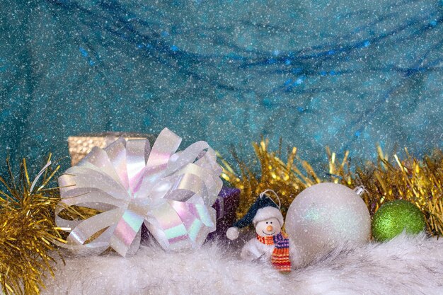 Un muñeco de nieve sonriente sobre pelaje blanco junto a bolas de Navidad y un gran lazo blanco. Fondo azul con el efecto de la nieve que cae. Tarjeta de Navidad. Enfoque selectivo en el muñeco de nieve.