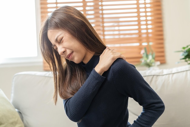 Músculos del cuerpo problema rígido asiático joven atractiva mujer niña dolor con dolor de espalda dolor de trabajo sosteniendo masaje frotando el hombro dolor o dolor doloroso sentado en el sofá en casa Personas de la salud