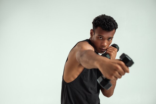 Músculos perfectos. Hombre africano joven muscular que ejercita con pesas mientras está de pie contra el fondo gris. Tiro del estudio. Concepto de deporte