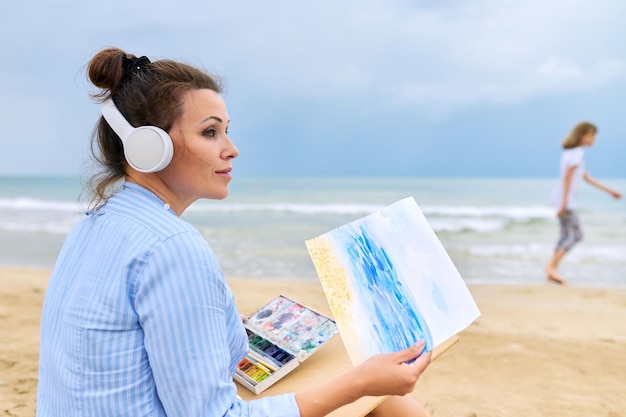 Música creatividad salud mental recreación al aire libre Mujer madura con auriculares escuchando música disfrutando de la pintura con acuarela sentado en la playa de arena