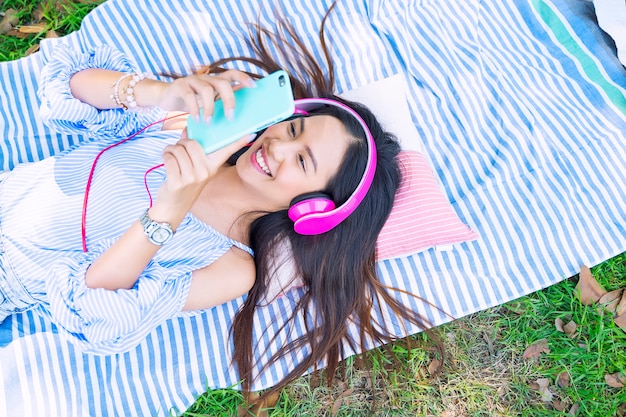 Música de escuta da jovem mulher com o smartphone e o fones de ouvido que encontram-se na terra no jardim com feliz.