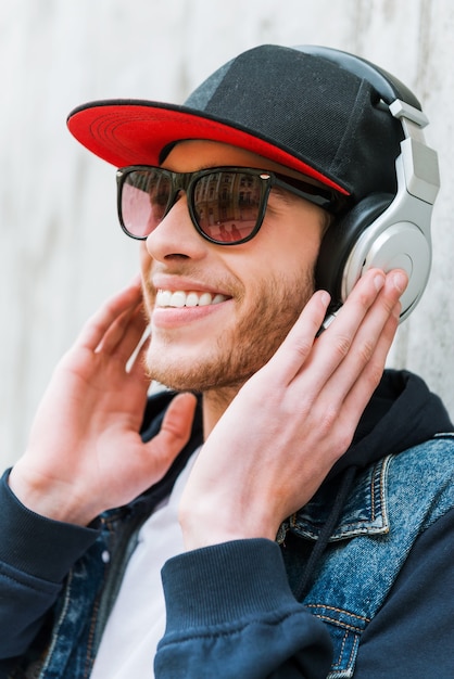 La musica es mi pasion. Feliz joven en auriculares sonriendo mientras se inclina contra la pared de hormigón