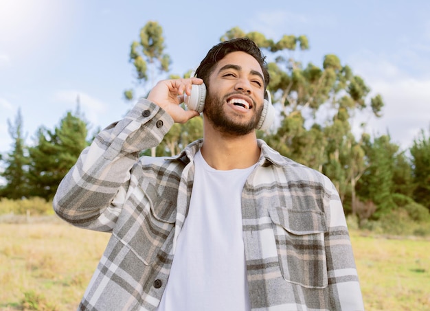 Música feliz e homem relaxando em um parque sorria e rindo de podcast ou faixa de áudio contra fundo da natureza Fones de ouvido riem e cara bonito ouvindo música de comédia online ou lista de reprodução na floresta