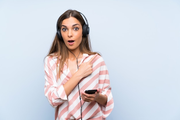 Música jovem com um celular sobre parede azul isolada surpreendeu e apontando o lado