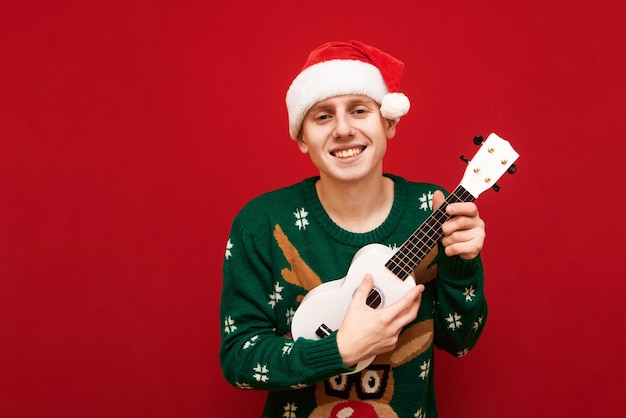 Músico alegre de retrato com chapéu de Papai Noel em fundo vermelho tocando ukulele