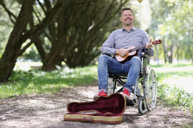 Músico callejero masculino toca la guitarra en silla de ruedas ganancias y beneficios sociales para discapacitados