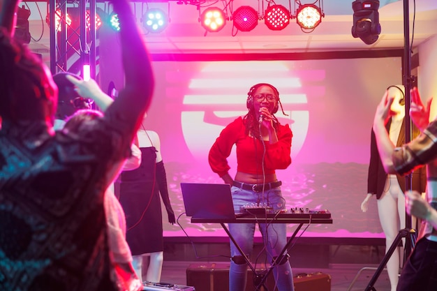 Músico cantando e tocando música eletrônica no palco de uma boate. DJ afro-americana usando microfone e equipamento de controle de som enquanto se apresenta em um clube lotado