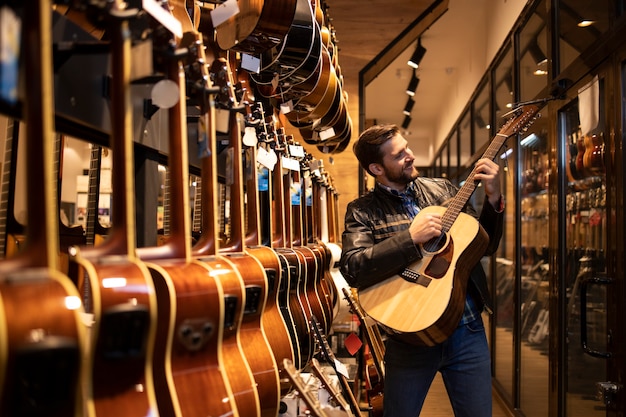 Músico caucásico talentoso en chaqueta de cuero comprobando el sonido del nuevo instrumento de guitarra en la tienda de música.