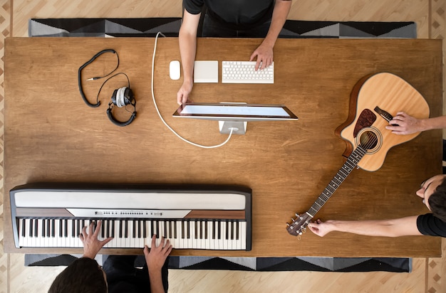 Músico creando música en su estudio tocando un teclado musical y una guitarra acústica. Proceso de trabajo en sonido en mesa de madera.