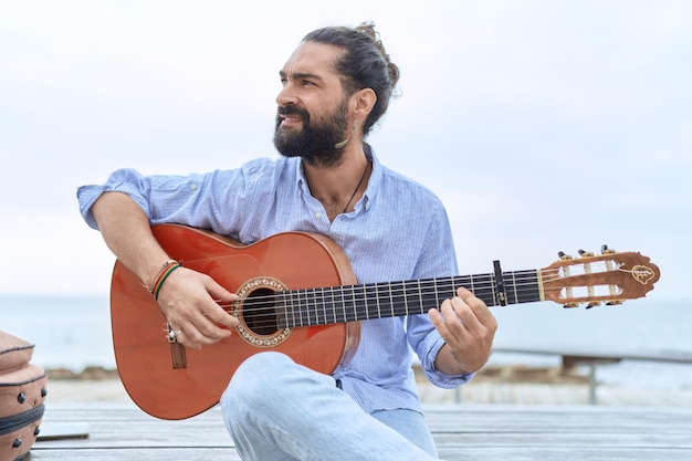 Músico jovem hispânico tocando violão clássico sentado no banco à beira-mar